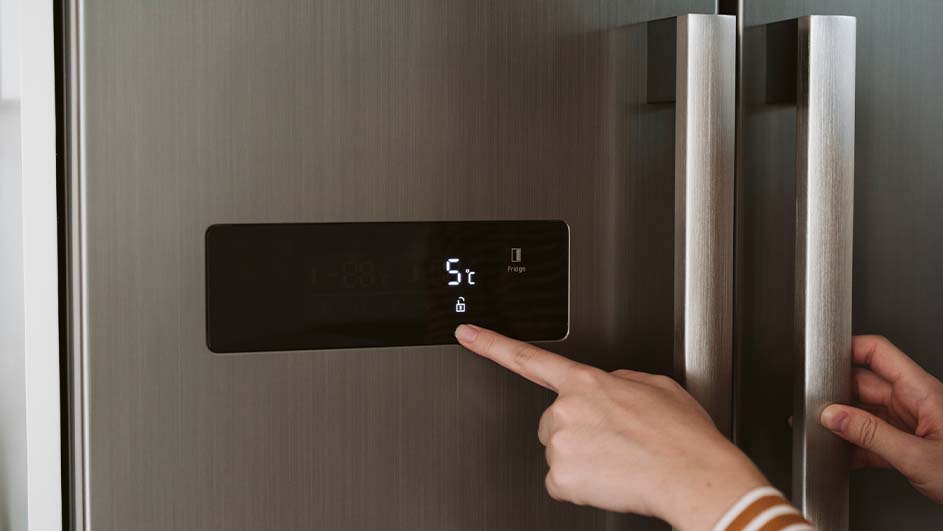 Een hand drukt op het display van een grijze koelkast met een temperatuur van 5 graden Celsius en een vergrendelingssymbool.