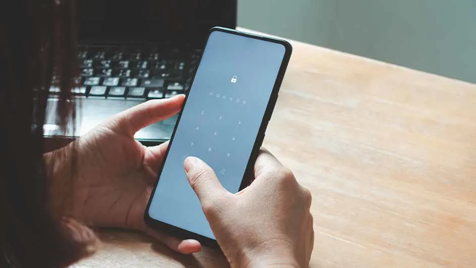Handen ontgrendelen een grijze smartphone met een numeriek slot, op een houten tafel naast een laptop.