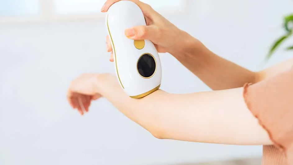 Een hand gebruikt een wit apparaat met gouden accenten op een arm in een lichte kamer met een plant op de achtergrond.