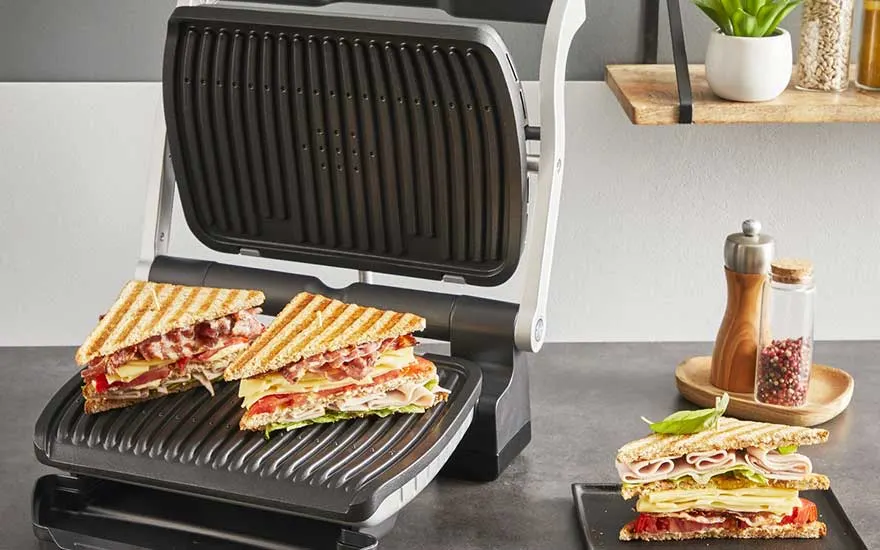 An open contact grill in a sleek kitchen toasts stacked club sandwiches, with extra fresh triangles on a plate ready to be served.