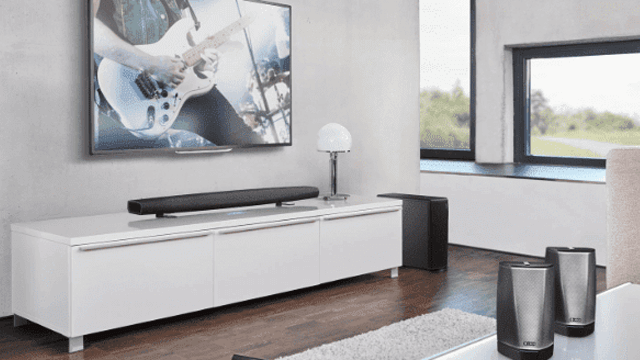 Woonkamer met tv waarop een gitaarspeler te zien is, witte kast met geluidsbalk, lamp, subwoofer en speakers.