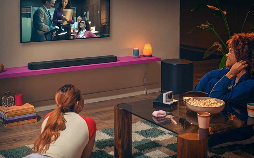 Twee vrouwen kijken tv in warme woonkamer met roze plank, soundbar, kom popcorn, kaars, plant en salontafel.