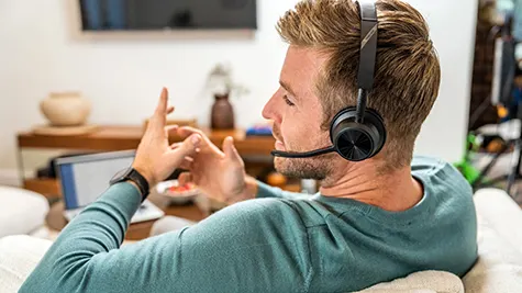 Man zit thuis op de bank met headset en micro, gebarend terwijl hij ontspannen deelneemt aan een online gesprek of vergadering.