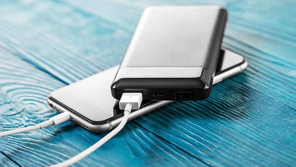 Een zilverkleurige telefoon wordt opgeladen door een zwarte powerbank op een blauwe houten achtergrond.