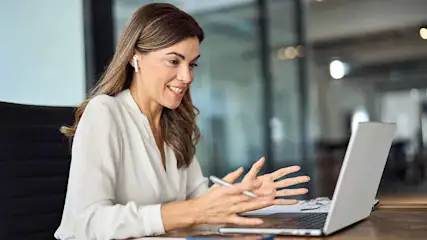 Vrouw met oortjes, witte blouse, en pen zit aan een bureau met een laptop in een kantooromgeving.