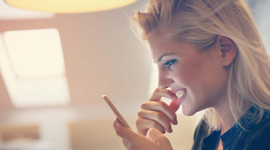 Een vrouw glimlacht terwijl ze naar een mobiele telefoon kijkt, met een hand aan haar mond en een raam op de achtergrond.