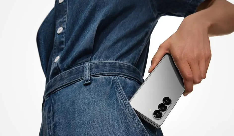 Een persoon in een denim outfit stopt een zilveren Samsung Galaxy Z Fold-smartphone met drie camera’s in de broekzak.
