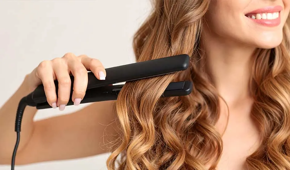 A smiling woman styles or shapes her long wavy hair with a black straightener against a light background.