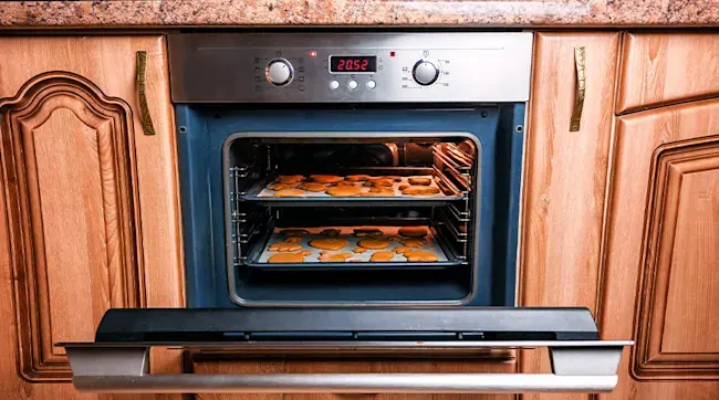 Cookies bake on two trays inside a blue oven, set in wood cabinets with metal handles. A digital clock reads 20:52.