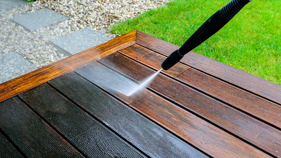 Een hogedrukreiniger die een houten terras schoonmaakt; het verschil tussen het schone en vuile oppervlak is duidelijk zichtbaar. Op de achtergrond zijn gras en grind te zien.