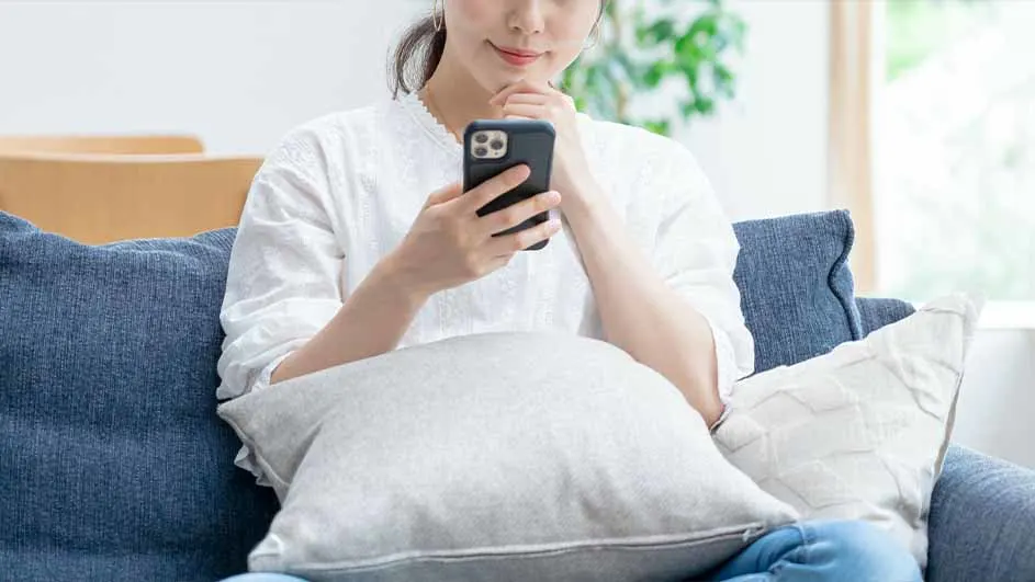 Vrouw zit op een blauwe bank met kussens, kijkend naar een zwarte smartphone. Er staat een plant bij het raam.