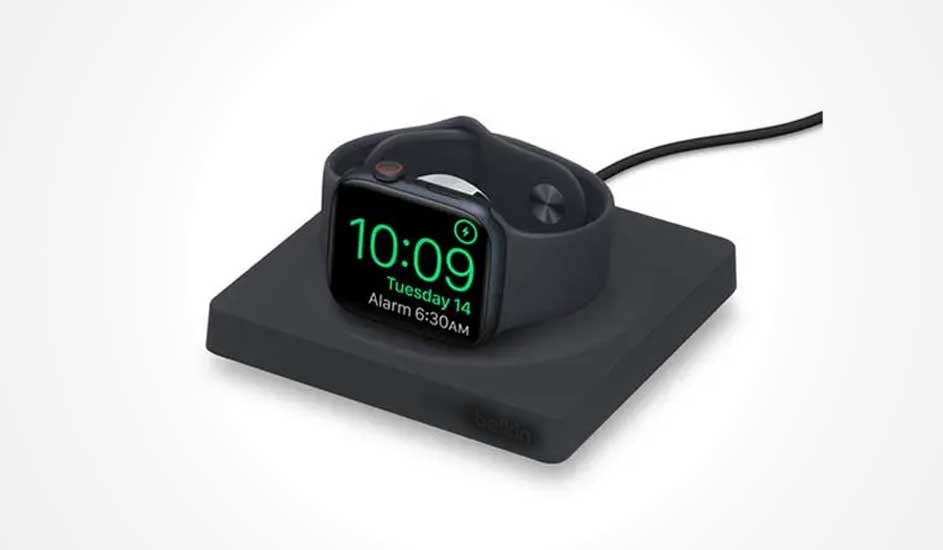 Black smartwatch resting on a dark charging pad, bright green digits display time, date and early alarm.