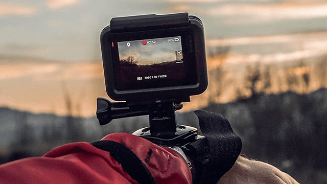 Een zwarte actiecamera filmt een landschap tegen een zonsondergang. De camera is bevestigd aan een rode mouw.