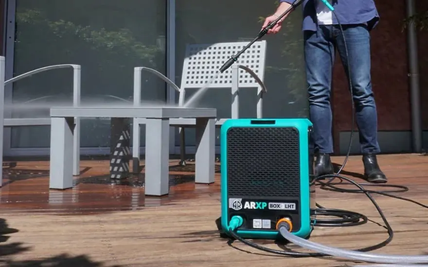 Iemand in spijkerbroek reinigt een houten terras met witte tuinstoelen, gebruikmakend van een turquoise hogedrukreiniger naast moderne meubels.
