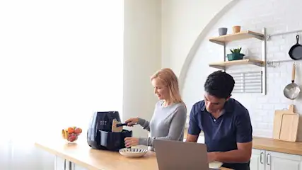 In een keuken staat een vrouw die eten in een zwarte airfryer plaatst, terwijl een man aan een laptop werkt.