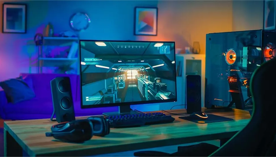A gaming setup features a computer, monitor displaying a game, keyboard, mouse, speakers, and headset on a wooden desk.