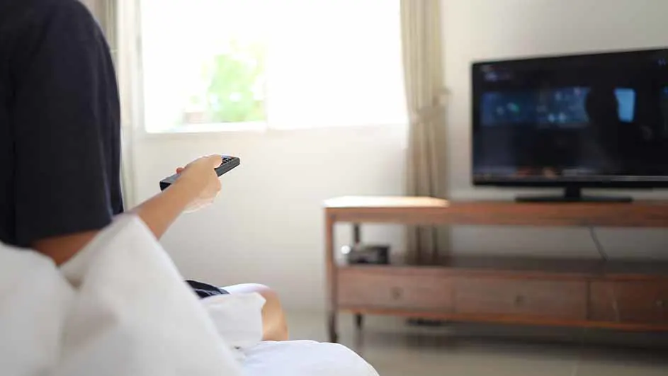 Zicht op iemand in bed met een afstandsbediening, een tv en een houten kast in een kamer met een raam.