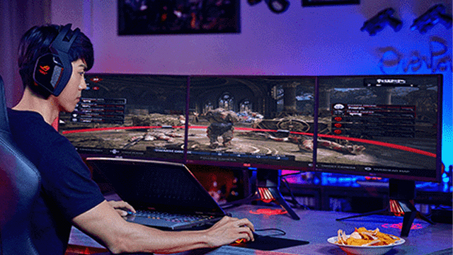 Een persoon met een koptelefoon speelt een computerspel op drie monitoren met een laptop en een schaal snacks.