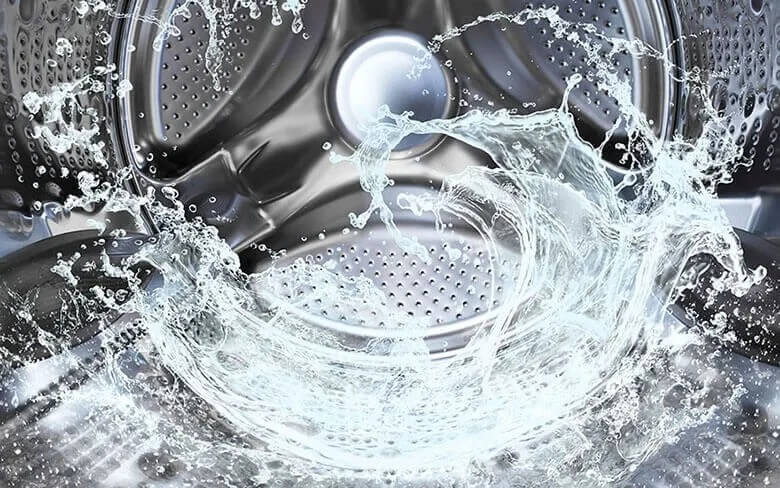 A dynamic image of water splashing inside a stainless steel washing machine drum. The perforations in the drum are clearly visible, and the water creates a swirling effect, suggesting motion.