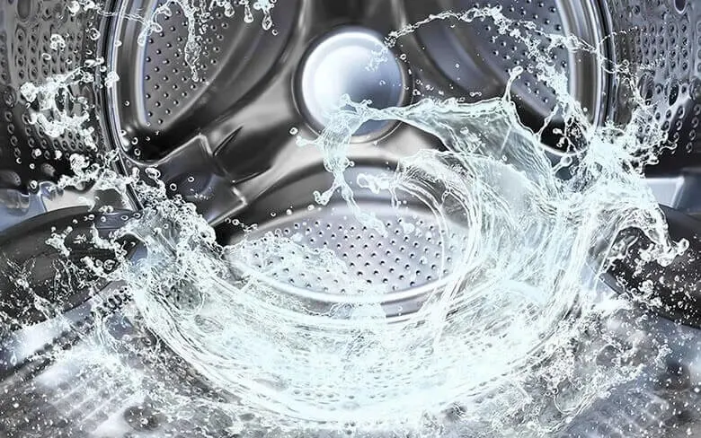 Een dynamisch beeld van opspattend water in een roestvrijstalen wasmachinetrommel. De perforaties in de trommel zijn duidelijk zichtbaar, en het water creëert een wervelend effect, wat beweging suggereert.