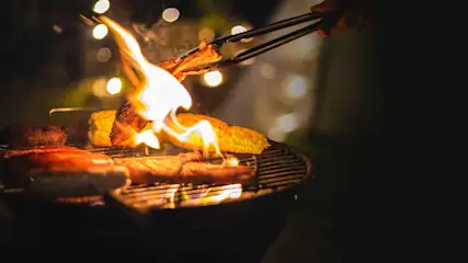 Een barbecue met maiskolven, vlees en vlammen. Een hand gebruikt een tang om het vlees te keren. Achtergrond met lichtjes.