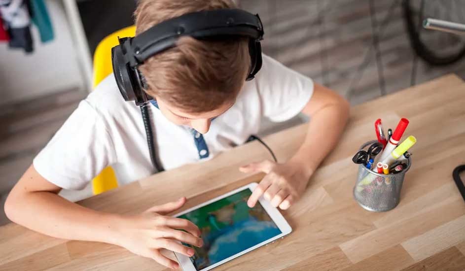A child wearing large headphones focuses on a tablet game at a wooden desk, with colourful pens and scissors in a pot nearby