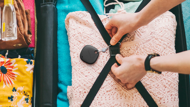 Een koffer gevuld met kledingstukken in diverse kleuren, zwarte riemen, een tracker en handen met een horloge.