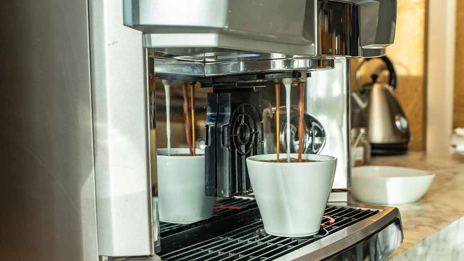 Coffee pours into two white cups from a stainless steel coffee machine on a kitchen counter with a kettle and bowl.