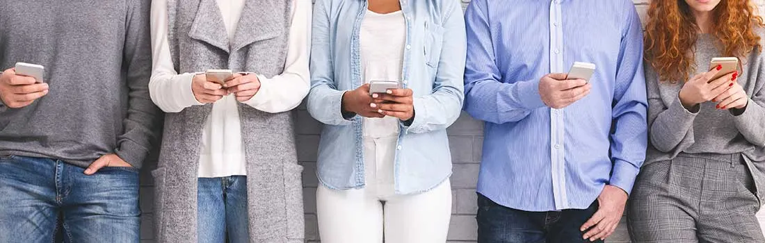 Vijf mensen staan tegen muur, elk met smartphone in handen, dragen casual kleding in grijs, blauw en wit.