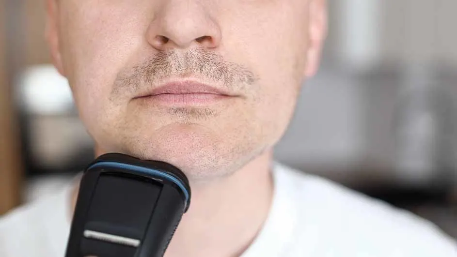 Een man gebruikt een zwart elektrisch scheerapparaat op zijn gezicht, vlak onder zijn lippen en op zijn kin. Hij draagt een wit shirt.