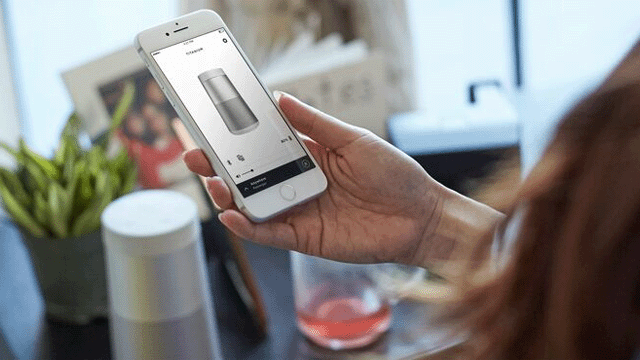 Een hand houdt een witte smartphone vast met een afbeelding van een speaker. Op tafel staat een speaker en een glas met rode vloeistof.