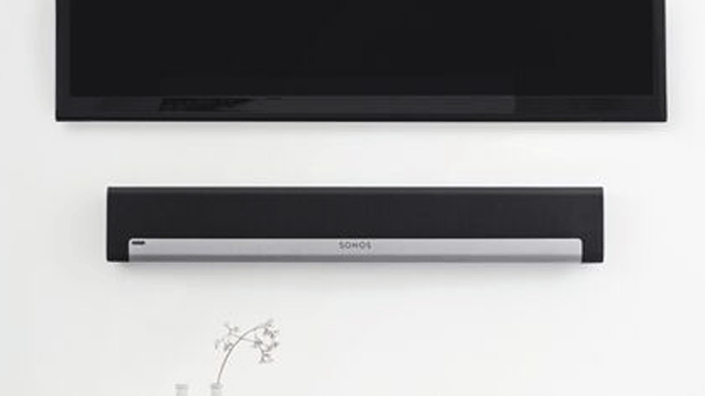 Zwarte televisie aan de muur boven een zwarte soundbar met zilveren accenten en een vaasje met bloemen.