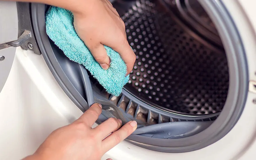 A person holds a blue towel to their face in a laundry room. A white washing machine, iron, and detergent sit on a shelf.