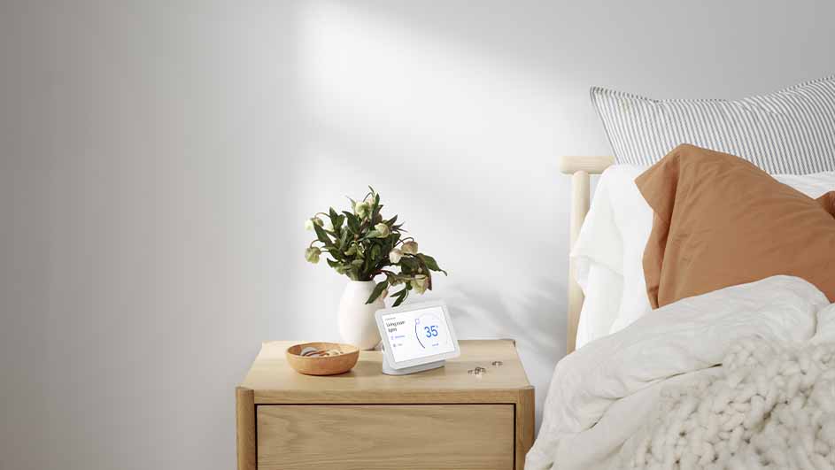 Bedroom scene showing a nightstand with a vase of flowers, a smart display, and a small bowl, next to a bed with pillows.