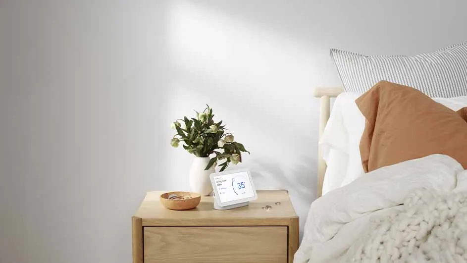Bedroom scene showing a nightstand with a vase of flowers, a smart display, and a small bowl, next to a bed with pillows.