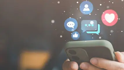 Handen houden een telefoon vast met digitale pictogrammen zoals een hart, chatballonnen en een persoon erboven.
