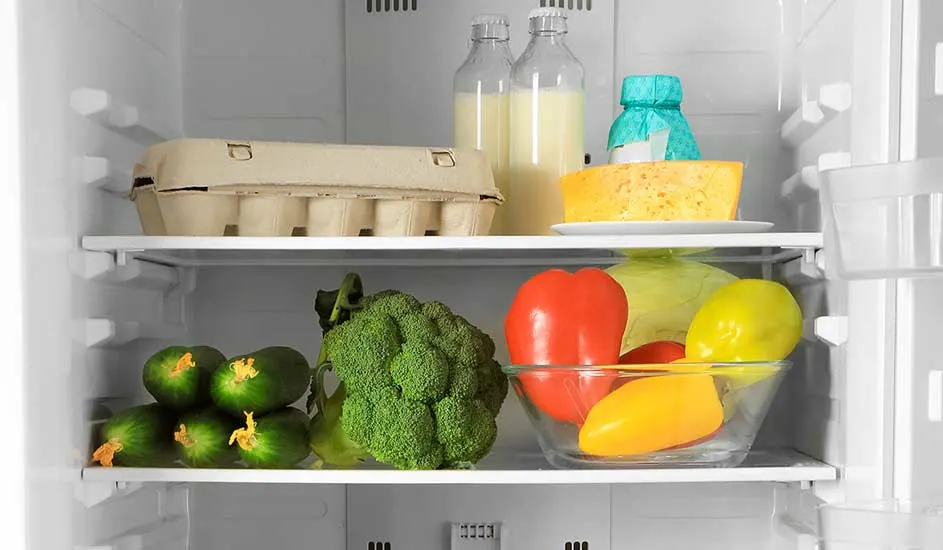 In de koelkast staan een doos eieren, twee flessen sap, een stuk kaas, komkommers, broccoli en een schaal met paprika’s en kool.
