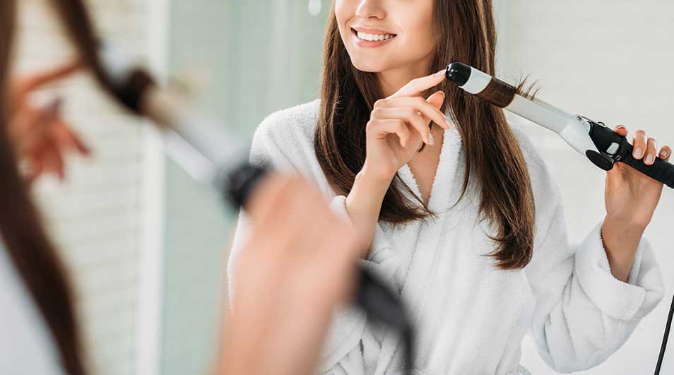A smiling woman in a dressing gown curls her hair with a curling iron; the background is blurred, as if someone watches.
