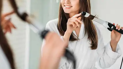 A smiling woman in a dressing gown curls her hair with a curling iron; the background is blurred, as if someone watches.