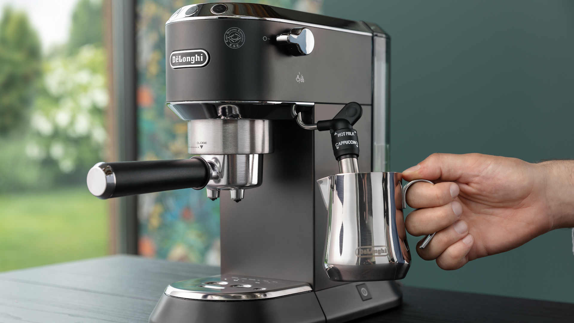 Een zwarte DeLonghi espressomachine staat op een tafel. Een hand houdt een stalen melkkan onder een stoompijpje.