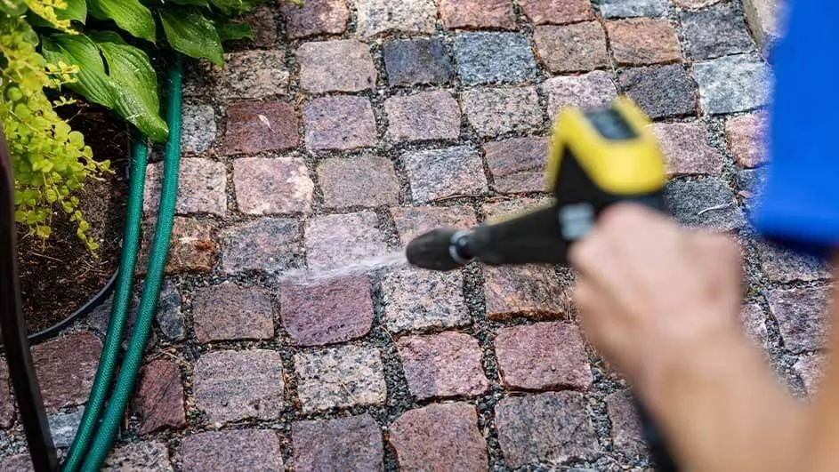 A Kärcher pressure washer is being used to clean a stone garden path, making the cobblestones visibly brighter near green plants.