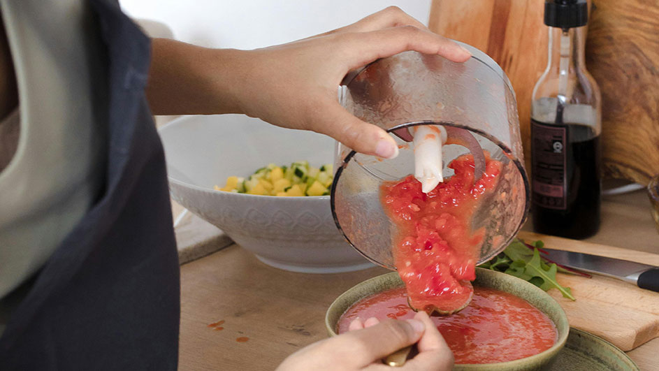 Iemand giet rode saus uit een blender in een groene kom. Op de achtergrond een salade, fles en houten plank.