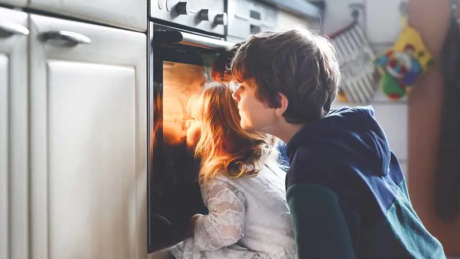 Twee kinderen kijken in een verlichte oven naast witte keukenkasten. Een kind draagt een blauw vest, het andere een witte jurk.