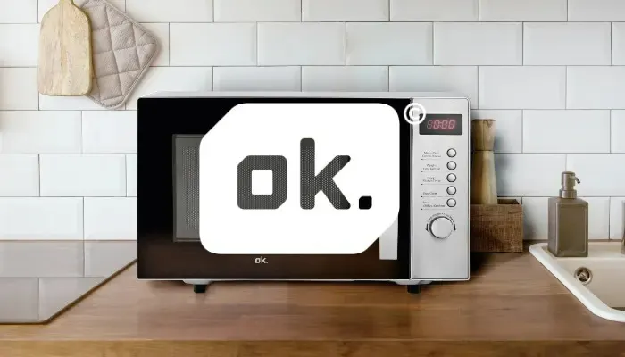 A microwave sits on a wooden counter, with a white tile backsplash. Oven mitts hang above. A sink and soap dispenser are visible.