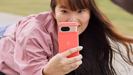 Een vrouw houdt een roze telefoon vast met een gouden camera, terwijl ze een roze jas en blauwe jeans draagt op een groene achtergrond.