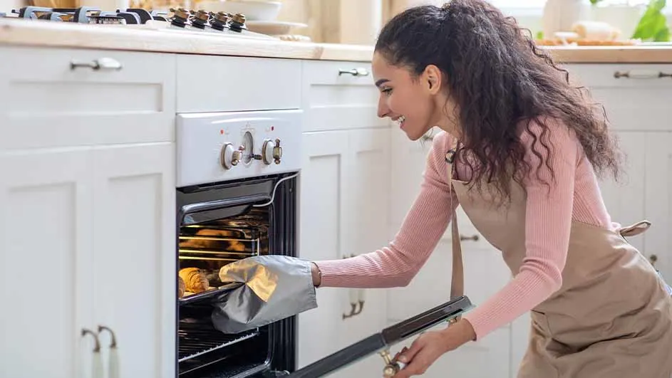 Een vrouw opent een witte oven met een ovenwant, terwijl ze in een lichte keuken staat.