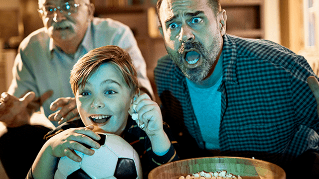 Drie mannen kijken naar een voetbalwedstrijd. Een jongen houdt een voetbal vast, een man heeft popcorn en een man is verrast.