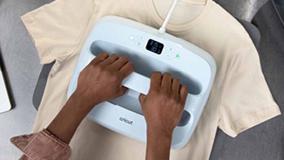 Twee handen houden een blauw Cricut-hittepers op een beige T-shirt. Een laptop staat op een grijze achtergrond.