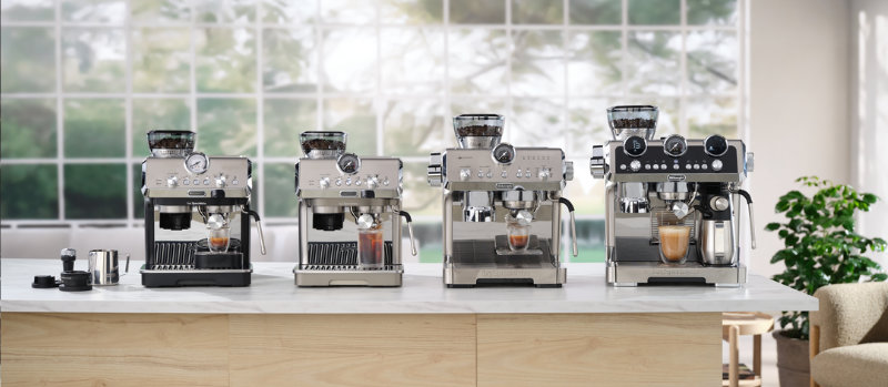 Vier zilverkleurige espressomachines staan op een aanrecht, met een groot raam en een plant op de achtergrond.