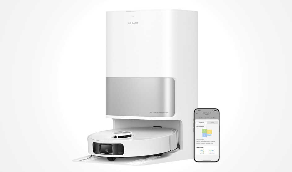 Een robotstofzuiger staat bij een bureau in een lichte woonkamer, met witte bank, radiator, raam en accessoires op een wandplank.